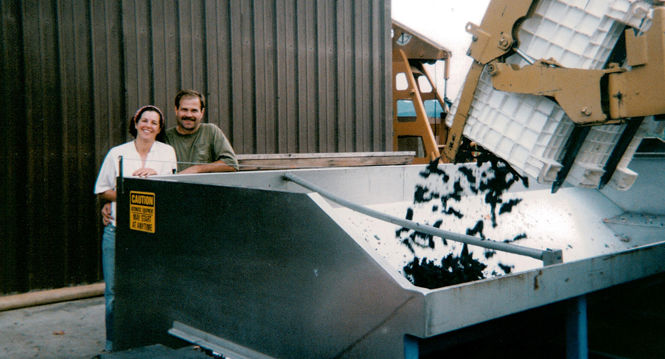 Two people stand next to a large metal container as a machine pours black grapes into it. The man on the right rests his arm on the edge, while the woman stands to his left. Both are smiling and dressed casually. The backdrop includes a corrugated metal building and machinery.