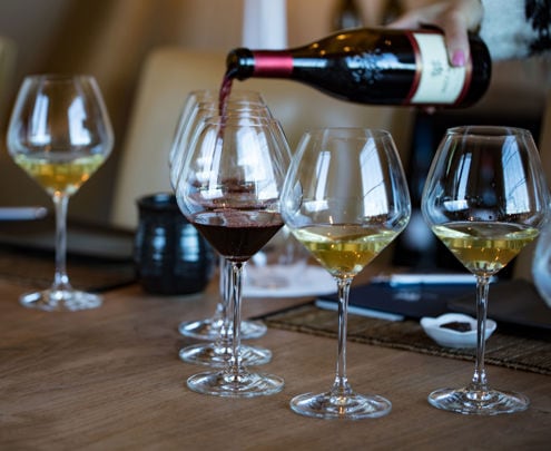 A person pours red wine from a bottle into one of five wine glasses arranged on a wooden table. The other four glasses are filled with white wine. The setting appears to be a cozy indoor space, possibly a dining room or wine tasting event, with neutral-toned decor and soft lighting.
