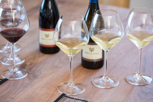 Three wine glasses filled with white wine and one glass with red wine are arranged on a wooden table. In the background, there are three wine bottles, each labeled "Brezi Hall." Sunlight softly illuminates the scene, highlighting the different shades of the wine.
