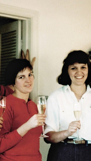 Four people are standing indoors, holding champagne glasses and smiling at the camera. They are in a well-lit space with a decorative plate on the wall in the background. The individuals are casually dressed. Two are wearing red tops and two are wearing white tops with one person also in blue.